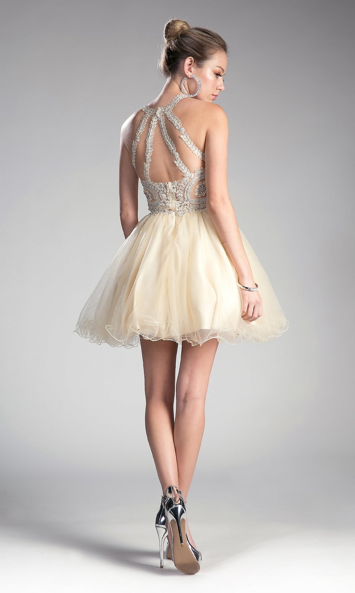 A woman stands against a neutral background wearing the affordable High-Neck Short Babydoll Homecoming Dress UJ0119, featuring an embroidered lace bodice, sleeveless and backless design, full cream skirt, and silver high heels.